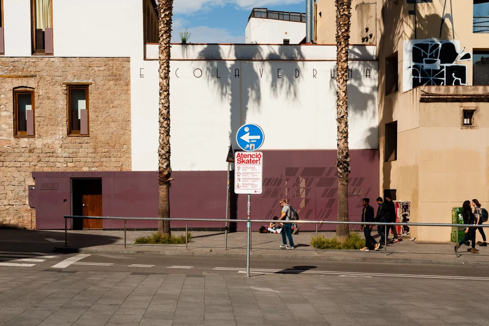 Urban scene with pedestrians on a sidewalk in front of modern buildings, palm trees, and street signage in bright daylight.