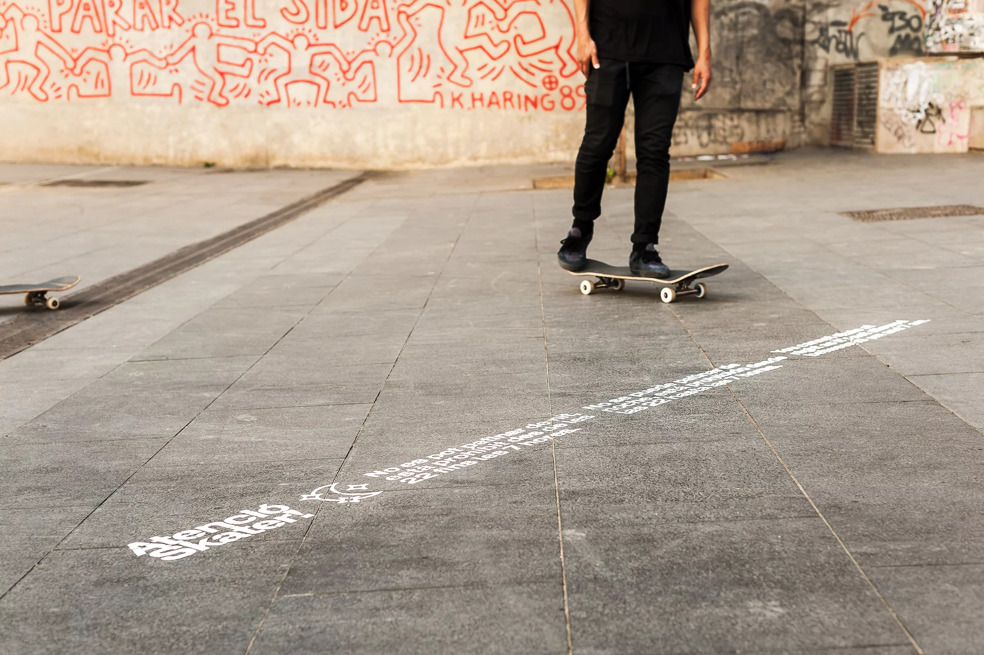 Skateboarder in black clothing rides on a skateboard in an outdoor urban area with graffiti art and text on the ground.
