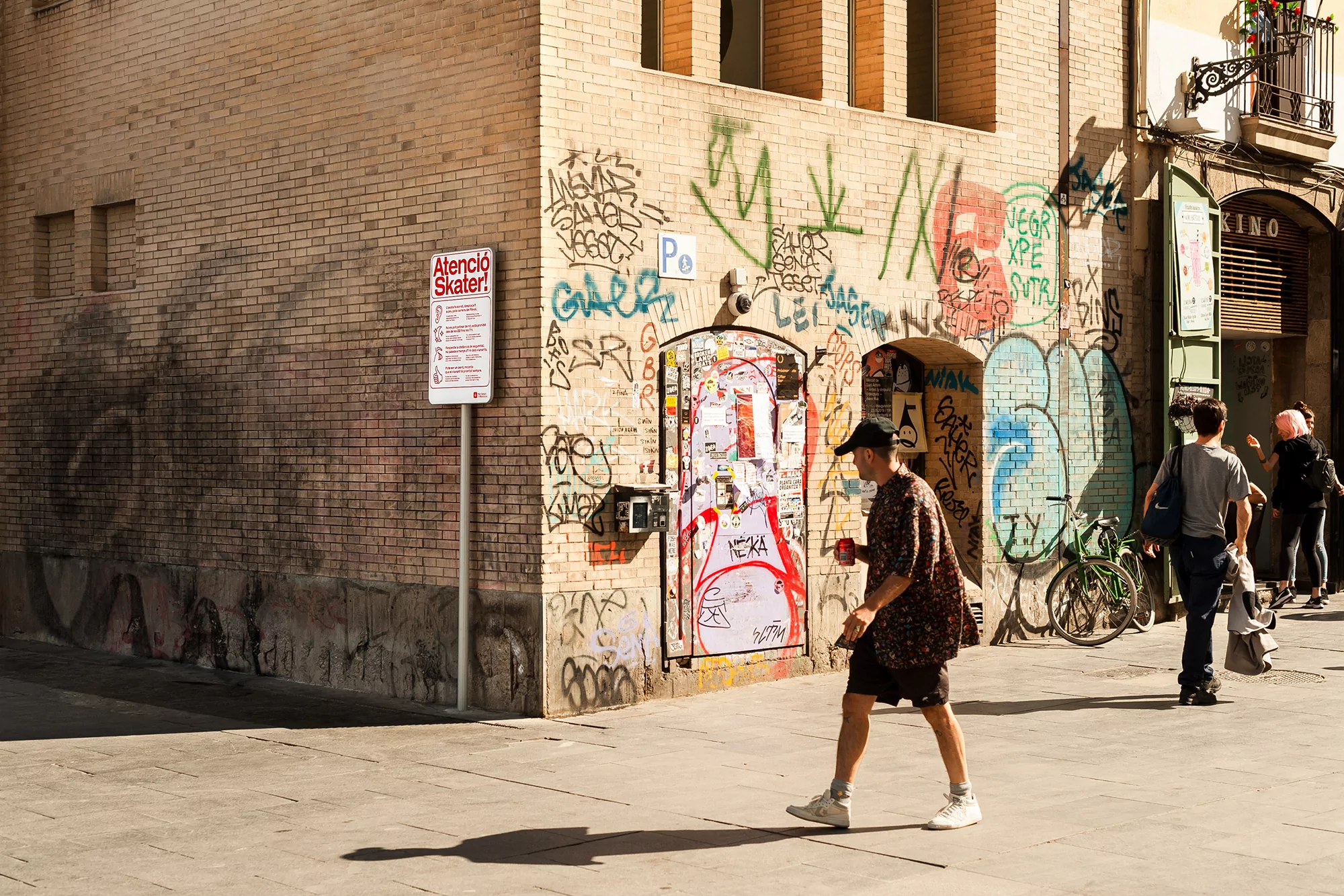 A man walks along a sunlit city street, with graffiti-covered walls and parked bicycles in the background, capturing an urban scene.