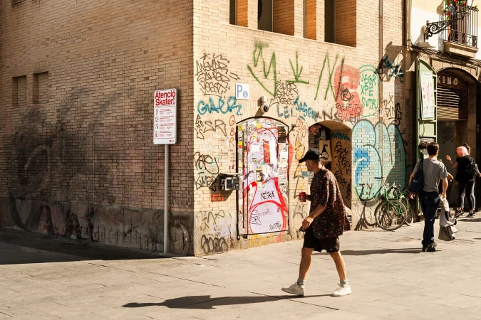 A man walks along a sunlit city street, with graffiti-covered walls and parked bicycles in the background, capturing an urban scene.