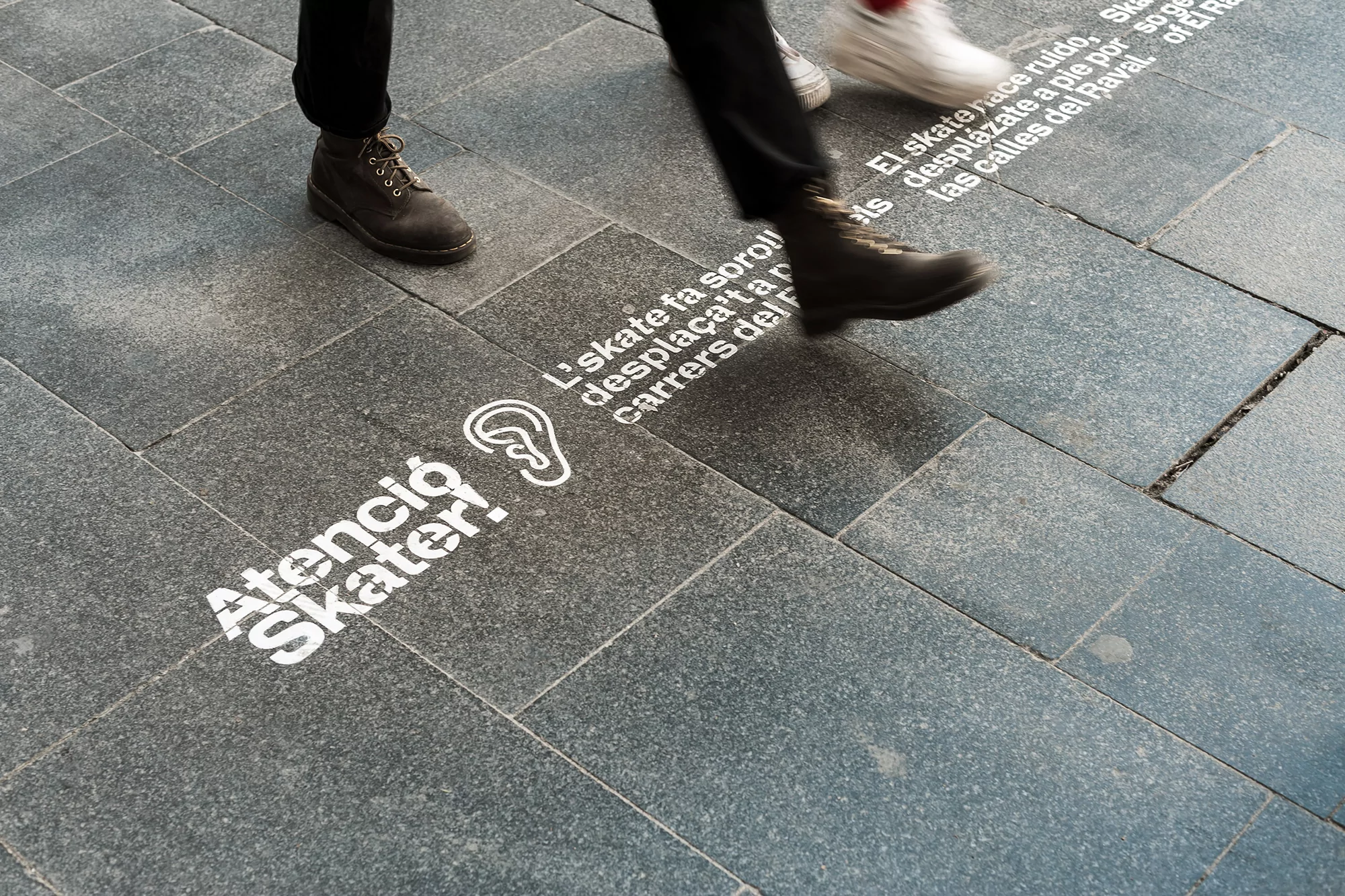 Two people walking on a city sidewalk with warning signs about skateboards painted on the pavement, boots and shoes in motion.
