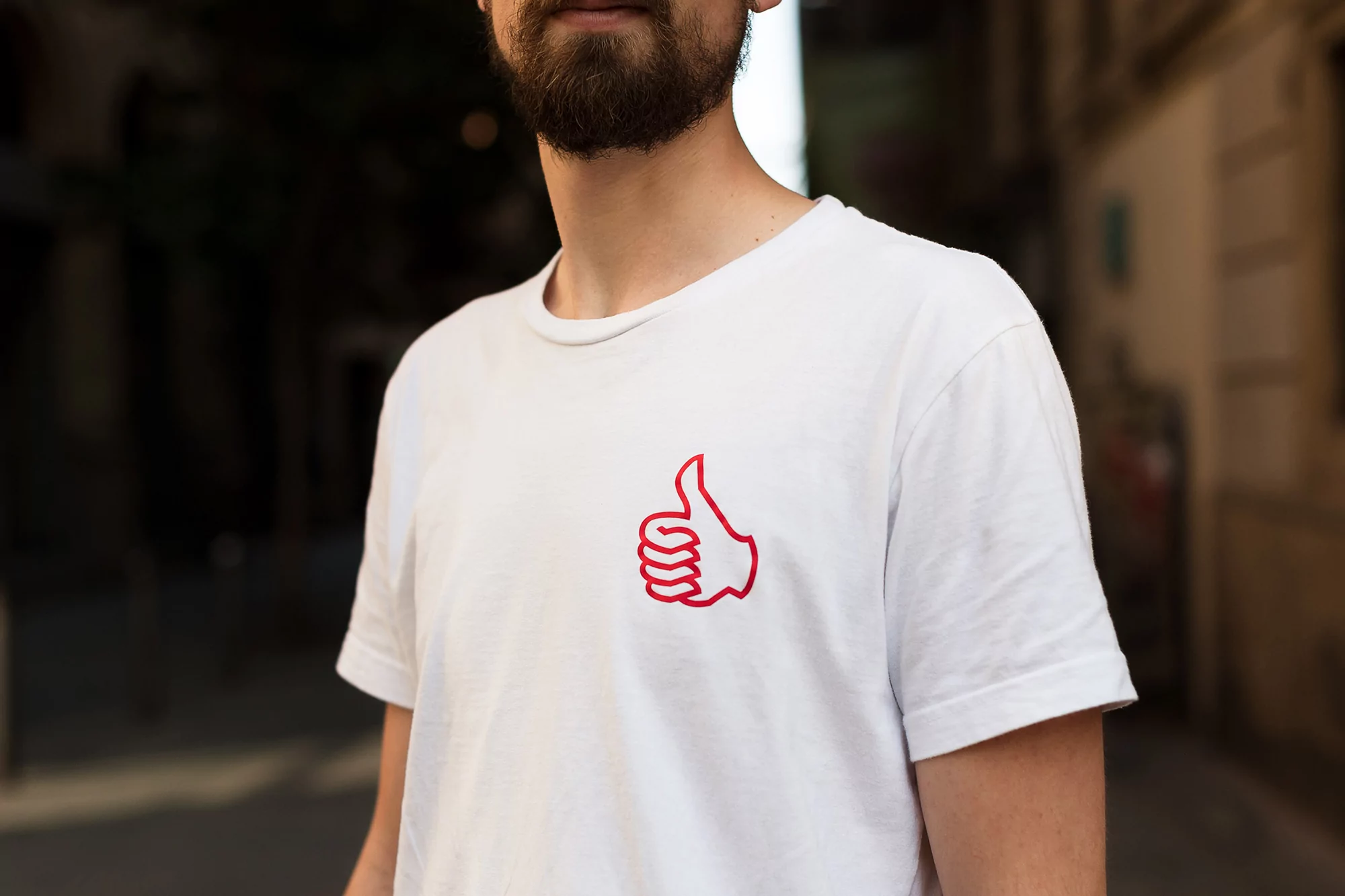 A man wears a white t-shirt with a red thumbs-up graphic, photographed outdoors in an urban setting with blurred background buildings.