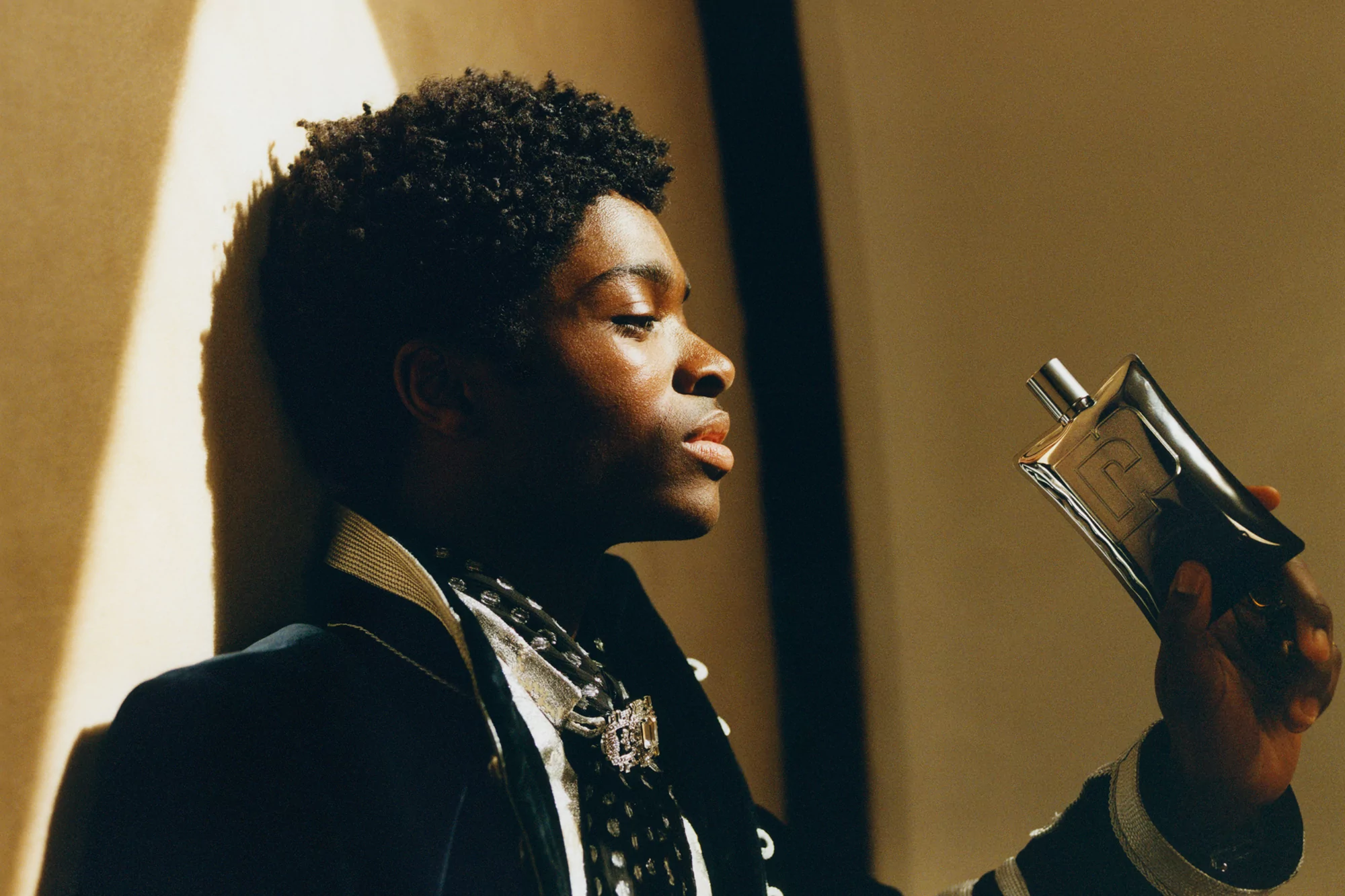 A black male model is posing indoors in a black jacket and scarf, while holding up a silver perfume bottle near a beige wall.