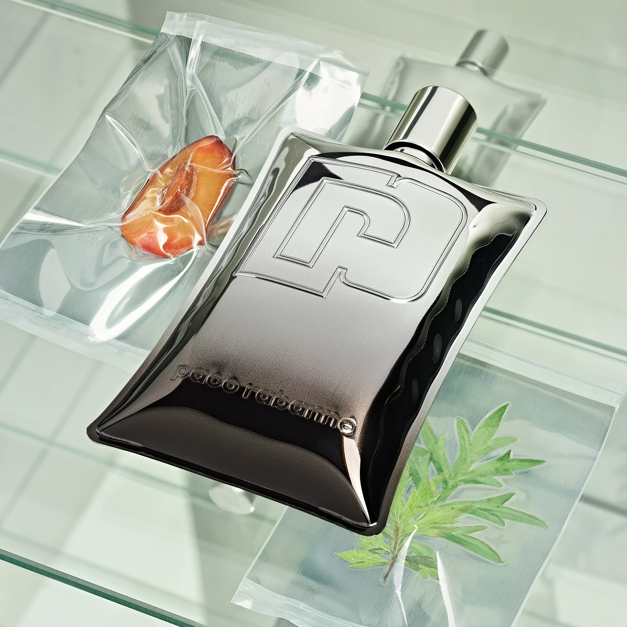 Studio shot of Paco Rabanne Phantom Parfum bottle, silver robot shape, surrounded by plants and meat in vacuum-sealed bags on glass.
