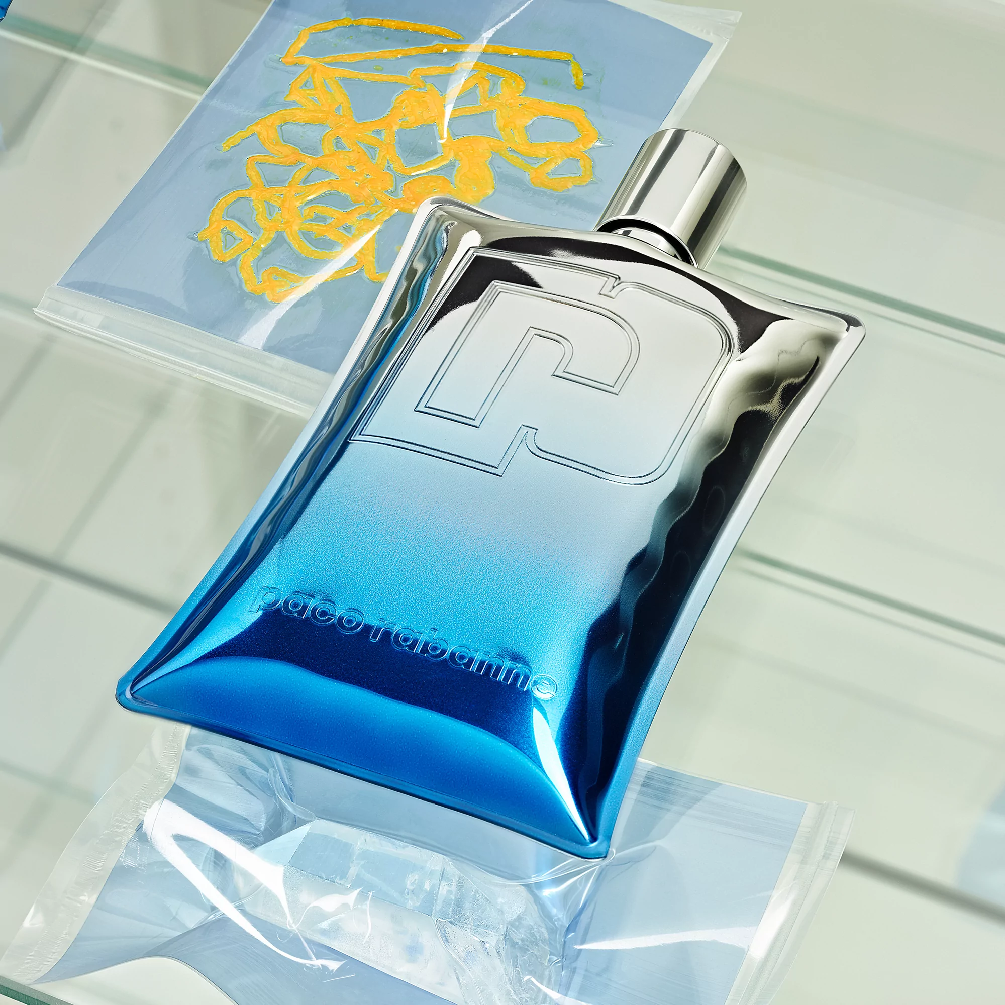 A shiny, partially transparent cologne bottle with a metallic silver top sits on a reflective glass surface with packaged art behind.