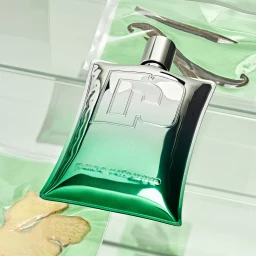 Metallic green Paco Rabanne perfume bottle resting on a reflective layered surface with packaged organic ingredients