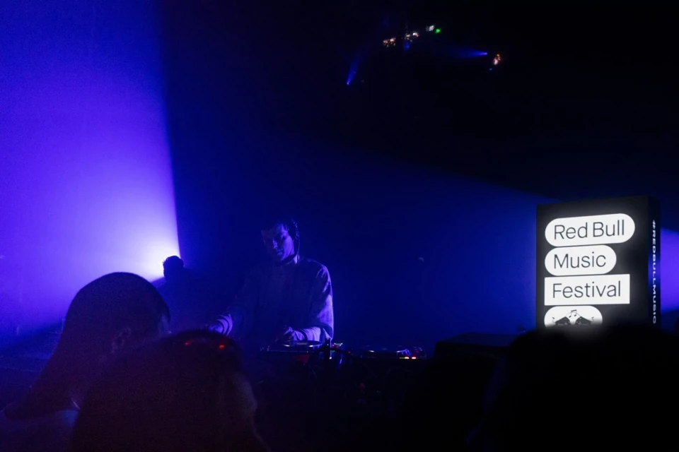 Live music festival scene with a DJ performing on stage bathed in blue light, crowd silhouettes, and a 'Red Bull Music Festival' sign.