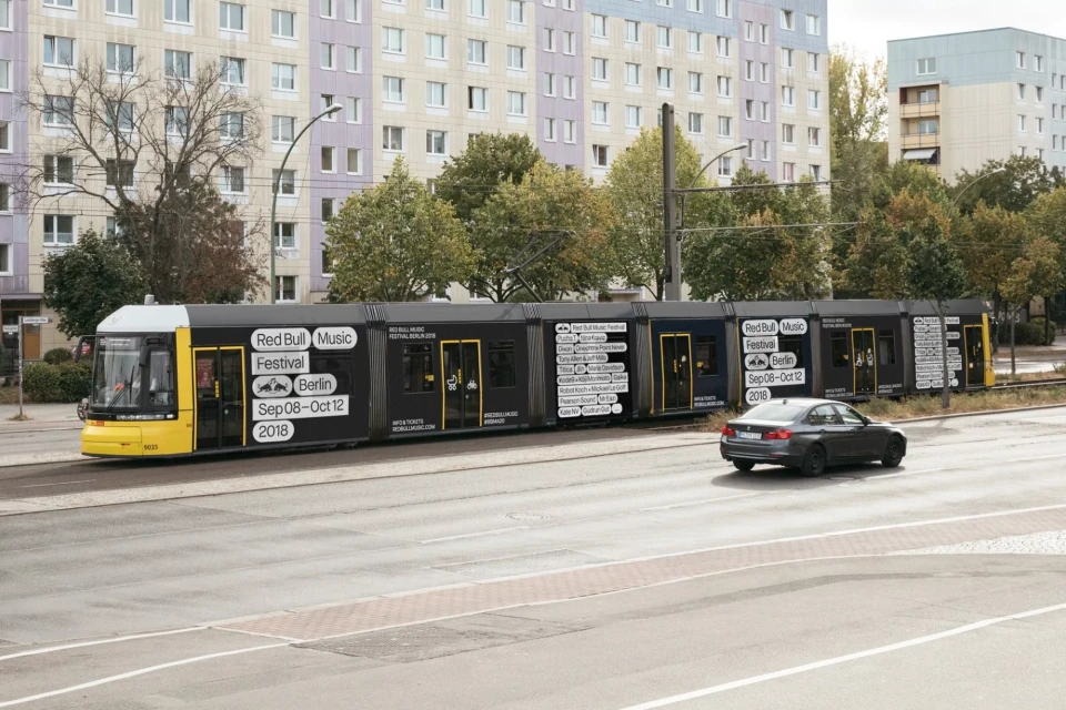 A yellow and black public transportation tram with Red Bull Music Festival Berlin advertisement, moving down a city street with a grey car.