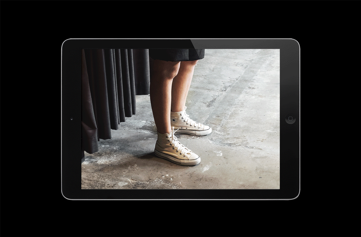Person wearing white high-top sneakers and a dark skirt, standing on a concrete surface, as seen on a black tablet device screen.