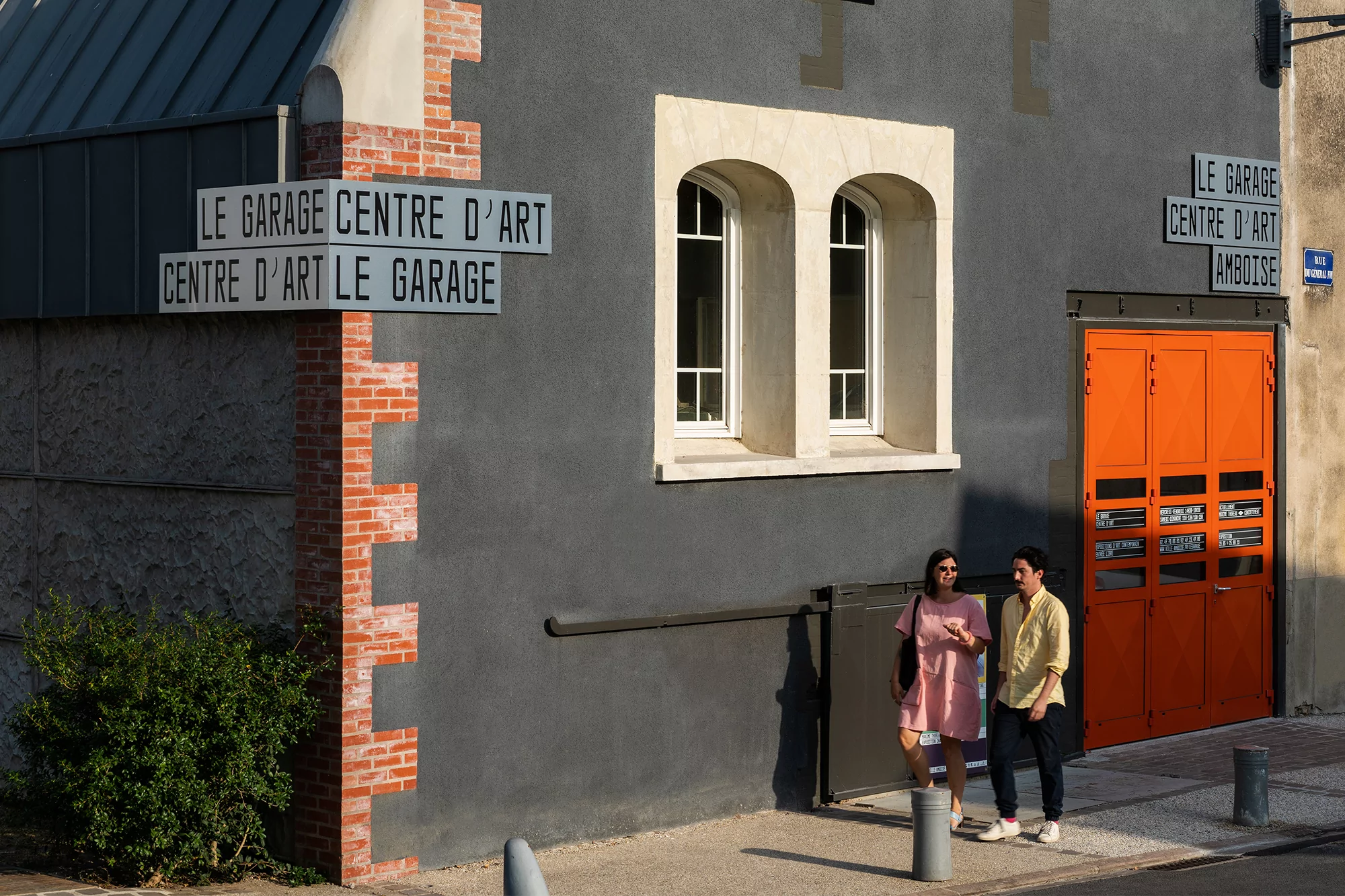 Two people stand outside Le Garage Centre D'Art, a grey building with an orange door and white-framed windows, under sunlight.