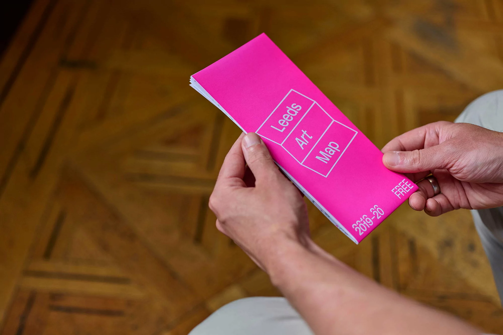 Man holding a bright pink Leeds Art Map booklet, showcasing a clean design against a blurred parquet wood floor background.