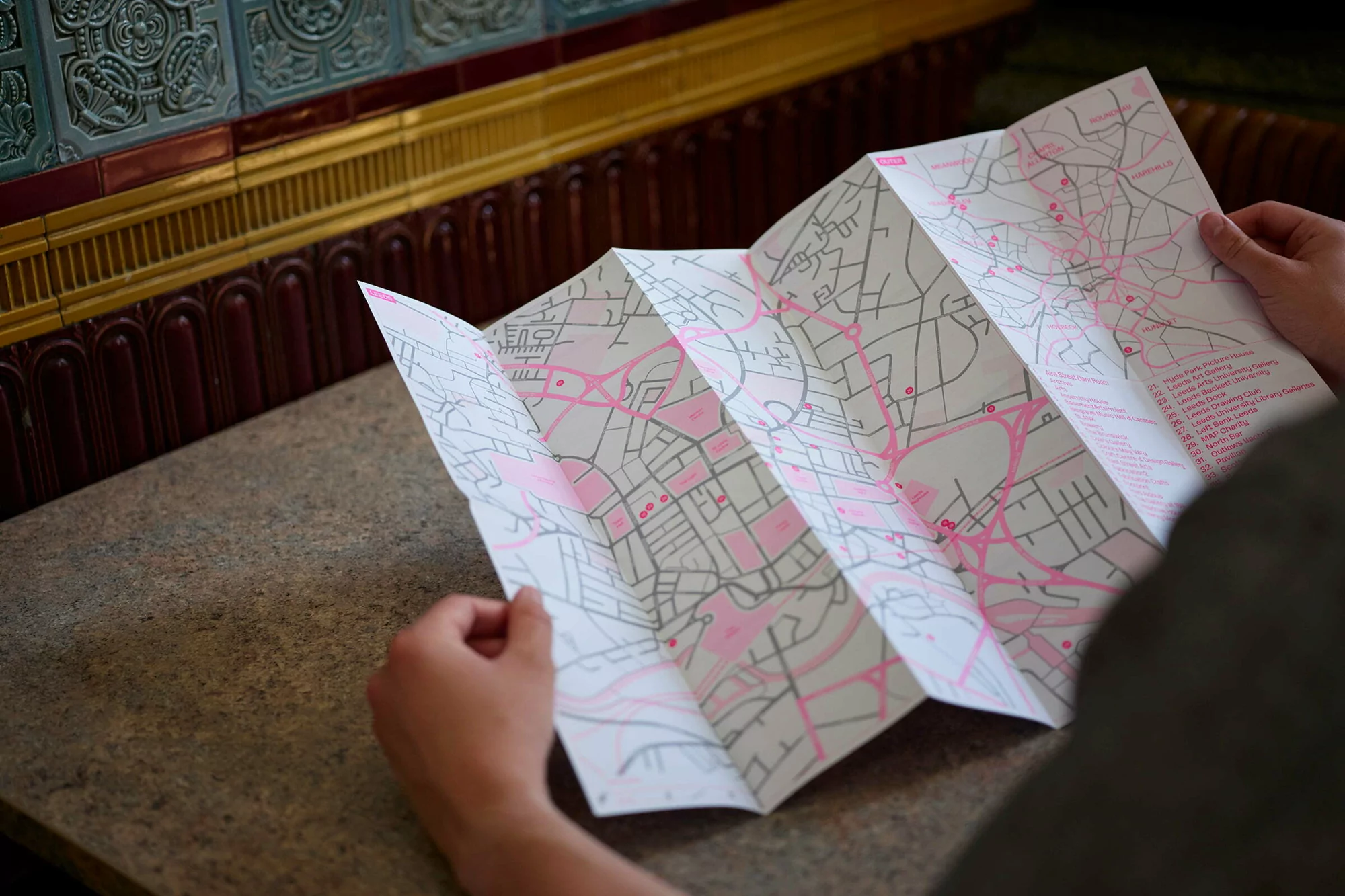 Close-up shot of an unrecognizable person holding an unfolded map, featuring pink route markings and text, against a tiled wall background.