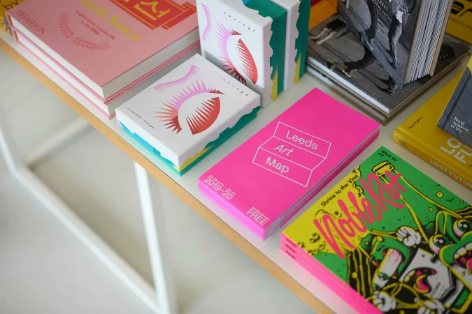 A high-angle view of colourful art books and zines arranged on a light wooden display table in a bright interior space.
