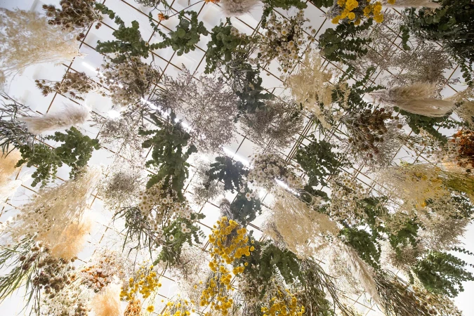 Dried flower arrangement suspended from a metal grid, featuring white, yellow, and green floral elements with a bright, upward perspective.