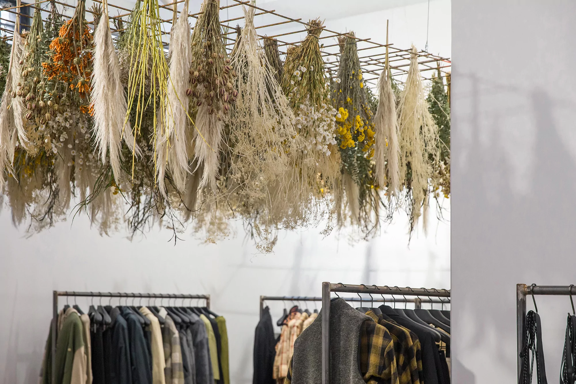 Dried floral arrangements hang above clothing racks in an indoor retail space. Garments on hangers include coats, jackets, and shirts.