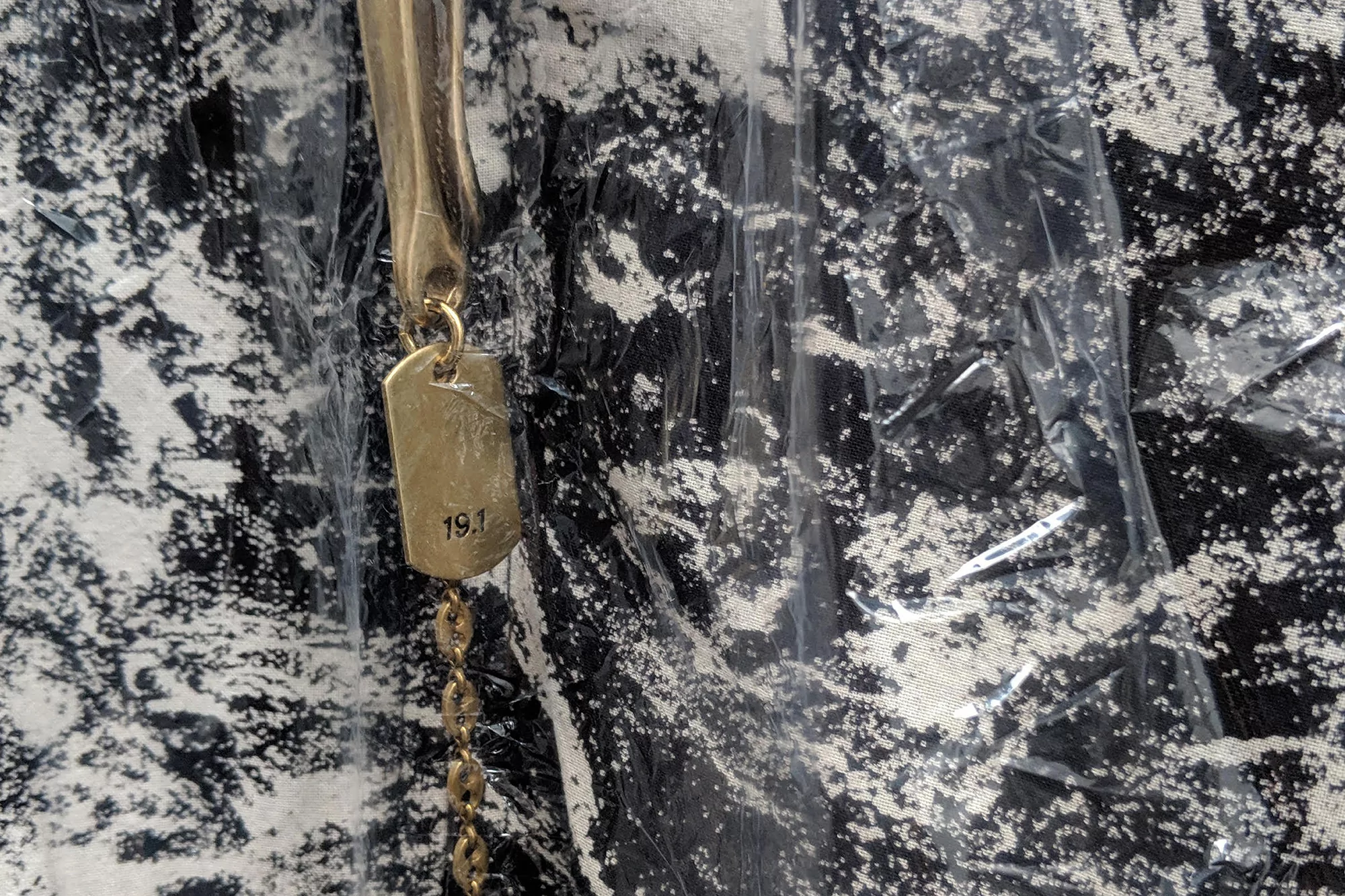 Gold-colored chain necklace with a tag and a black and white patterned bag enclosed in clear plastic packaging for protection.