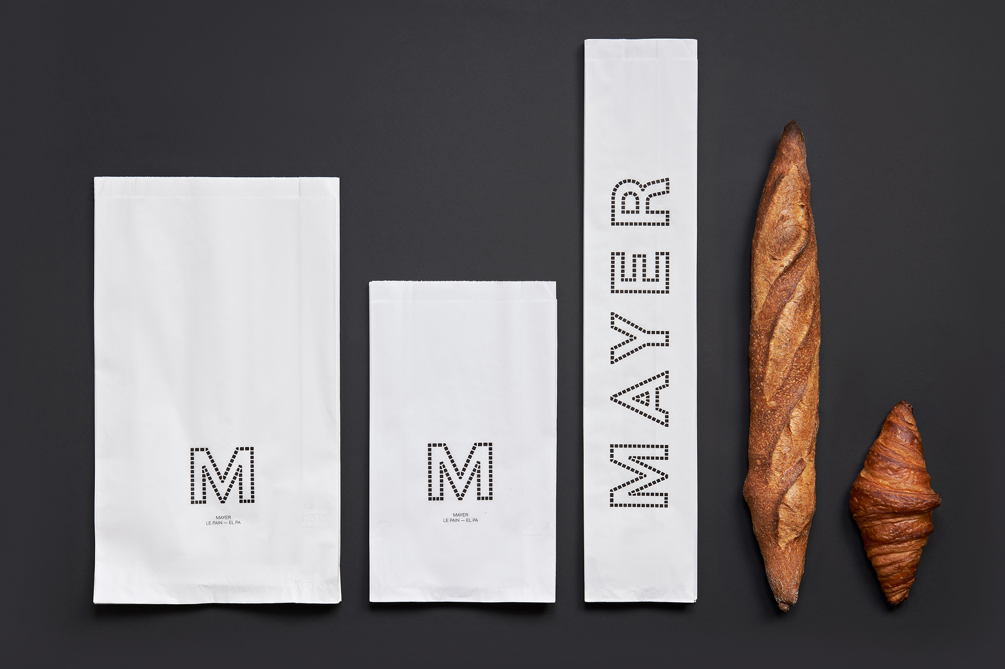 Artisanal baked goods presented with branded paper bags on dark grey background, showcasing baguette, croissant, and 'Mayer' logo.