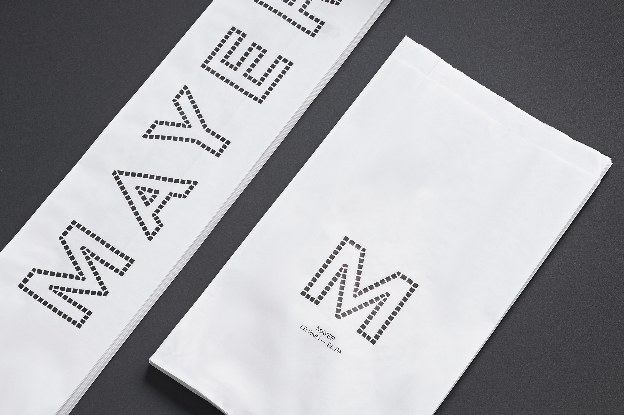 Close-up of stacked white paper bags with black lettering, featuring a minimalist design and arranged on a dark gray background.