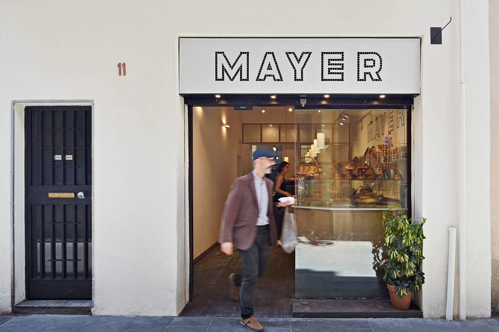 Man exits Mayer bakery store with a bag in an urban setting. The store has a large sign. Another person is visible inside.