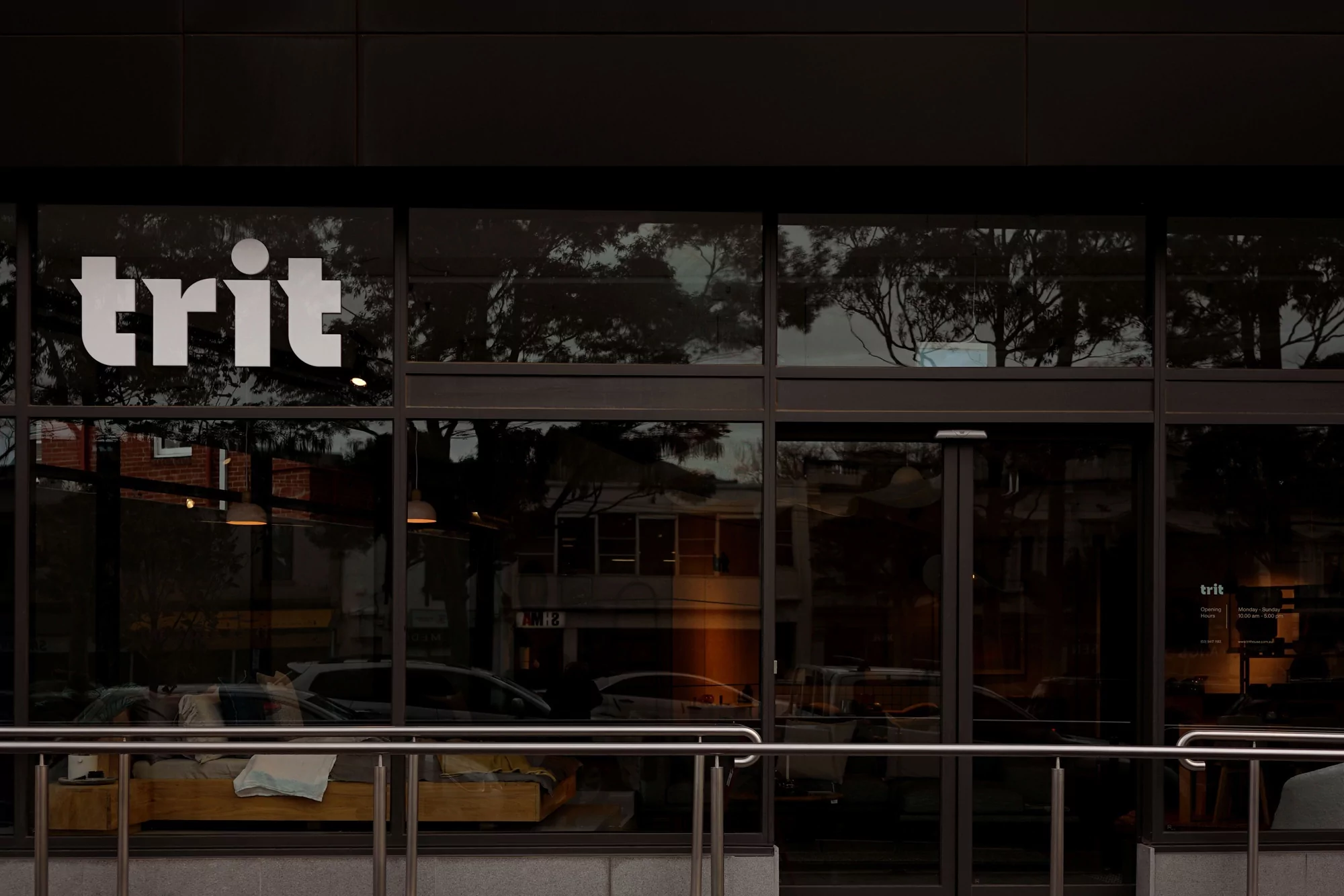 Exterior view of a commercial building with large windows displaying the 'trit' logo, reflecting outdoor trees.