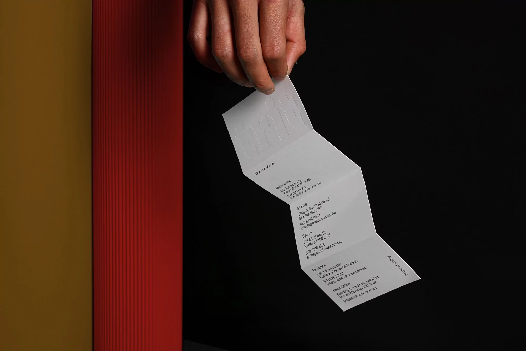 Hand holding folded white paper brochure against a black backdrop with red and yellow pillars
