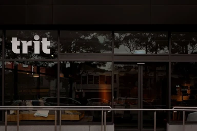 Storefront exterior of Trit featuring large glass windows with white brand typography and a metal railing in the foreground