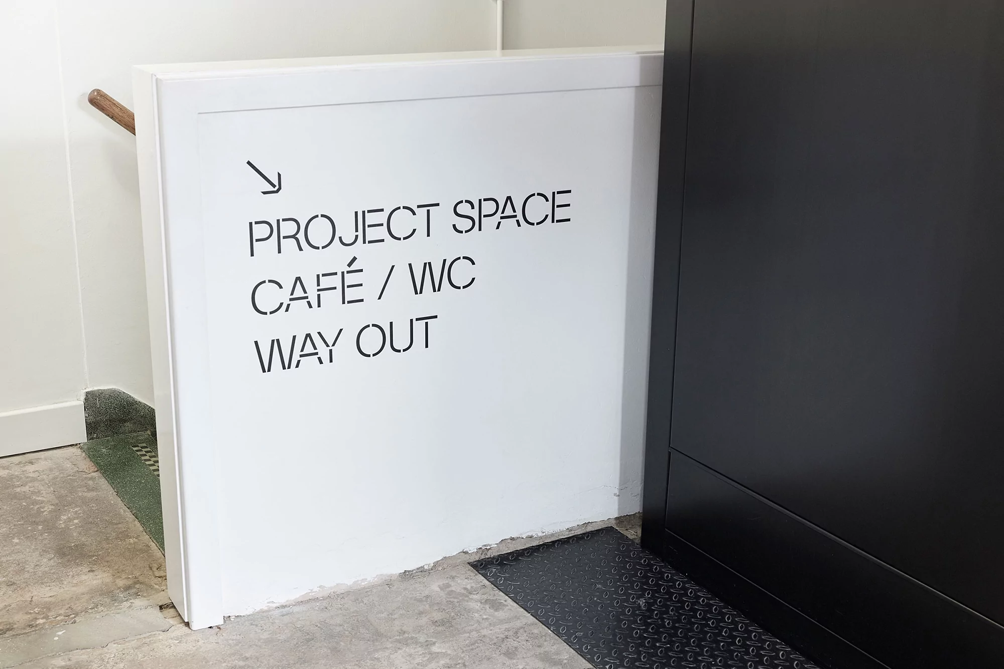 Directional sign with arrow pointing to project space, café, WC, and way out in a modern white hallway with a black element.