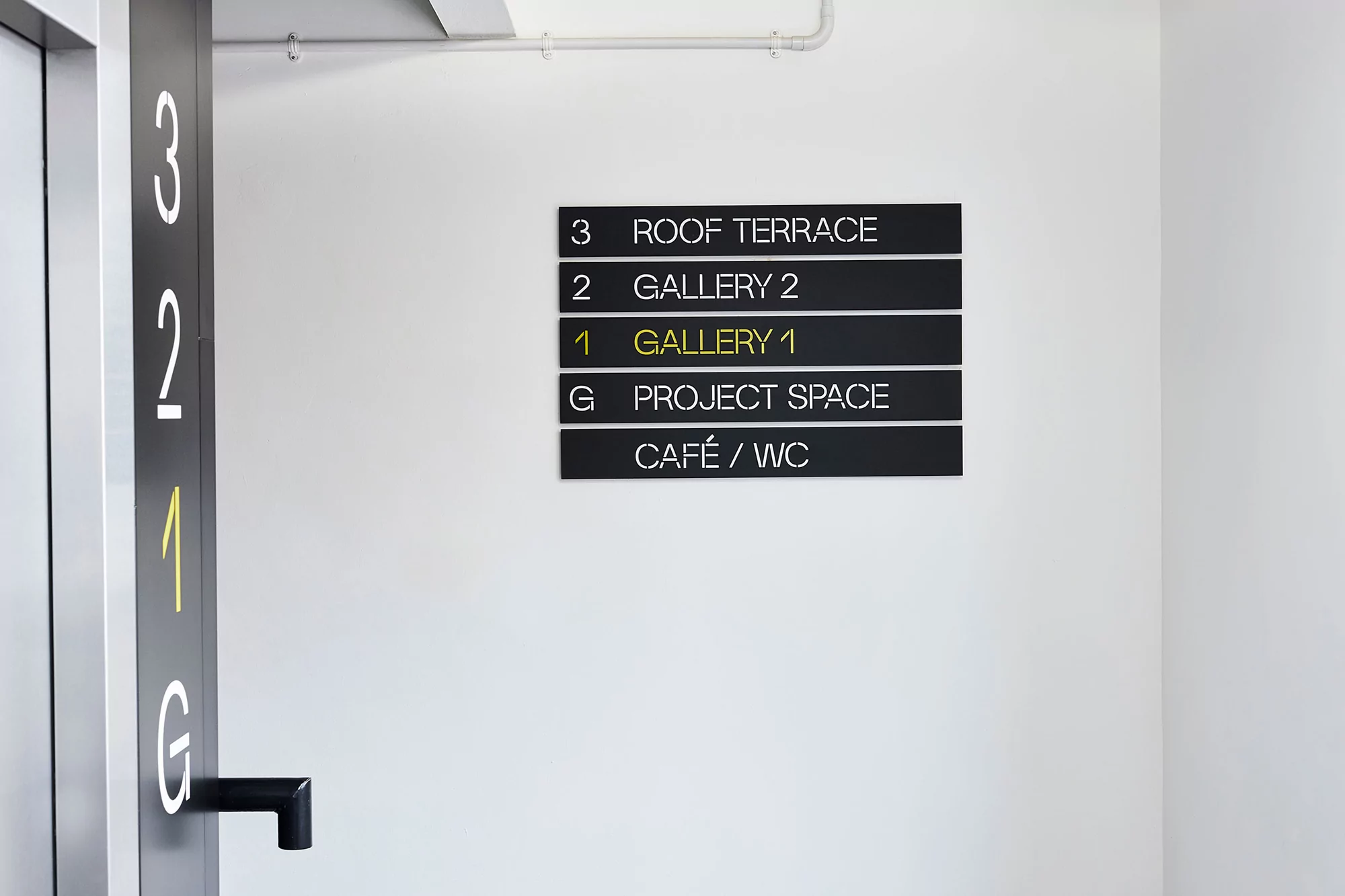 Interior wayfinding signage on a white wall directing to a roof terrace, galleries, project space, and café/WC on various floors.