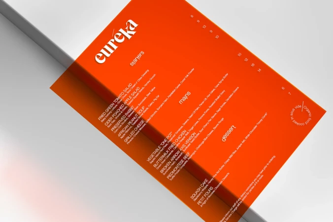 Close-up of an orange restaurant menu featuring starters, mains, and desserts, with the restaurant name 'Eureka' at the top.