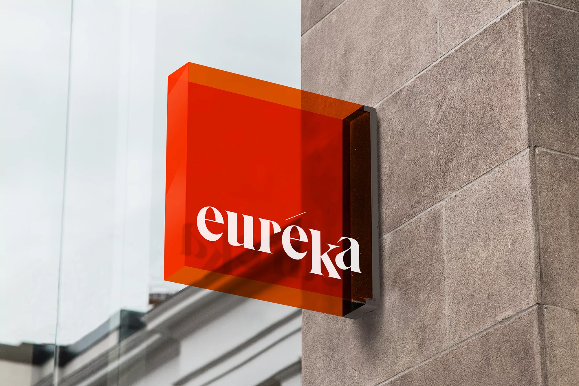 Exterior view of a square orange sign with the word 'eureka' in white letters mounted on a textured grey stone building facade.