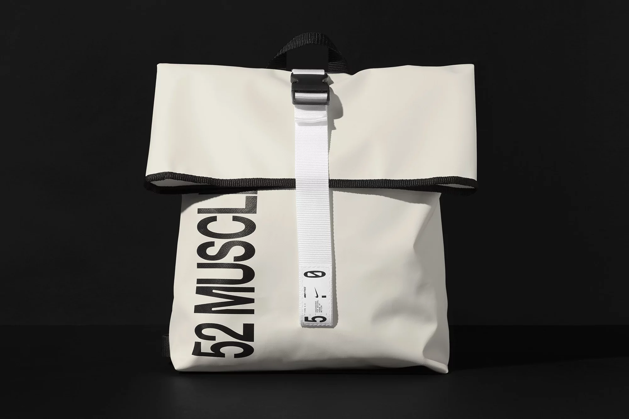 Cream-colored roll-top backpack featuring a white vertical strap and black block lettering isolated against a dark background.