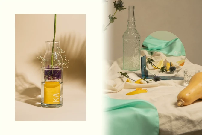 Still life composition featuring a glass bottle vase with flowers, a bottle, a mirror, cloth, and fruit on a beige background.