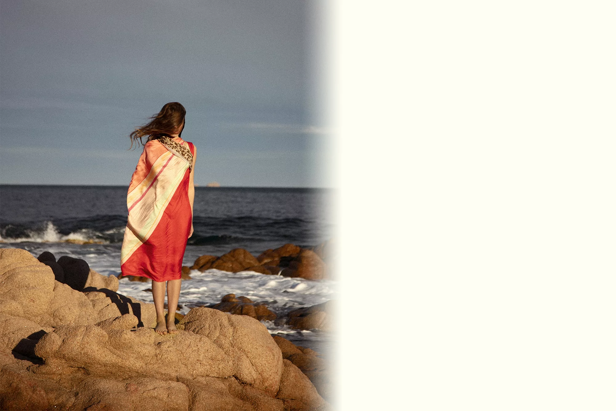 A solitary woman stands on weathered rocks facing the ocean, draped in a striped red and white fabric, wind gently blowing her hair.