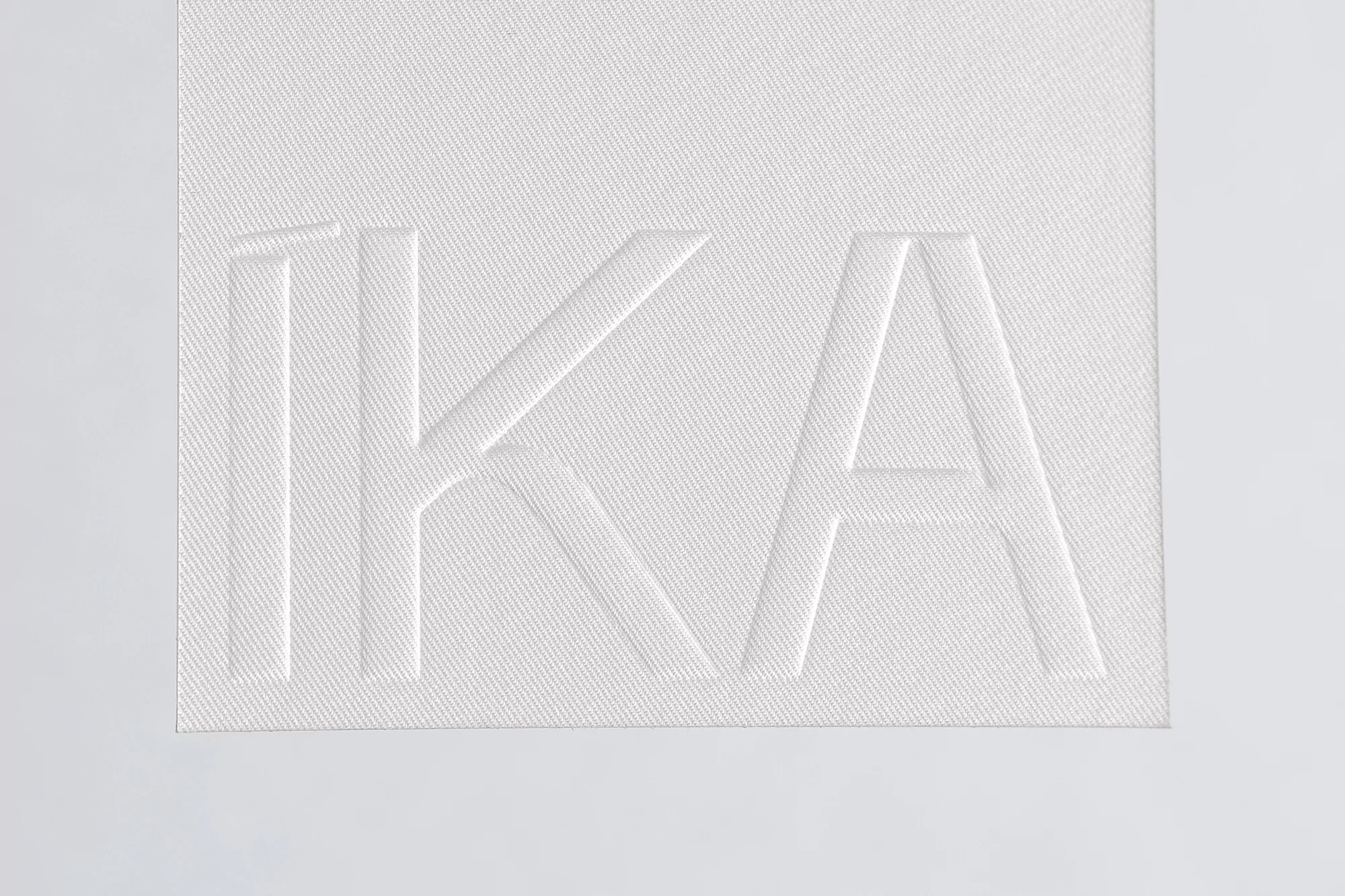 Close-up of textured white fabric with embossed 'IKA' lettering creating a minimalist, high-contrast abstract pattern and clean visual.