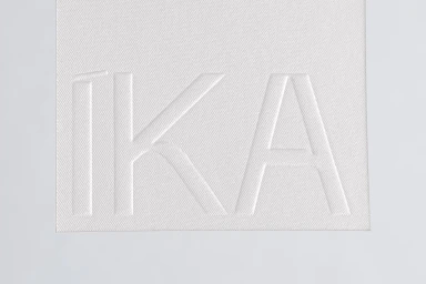 Embossed white paper brand identity featuring the name IKA on a textured white fabric background