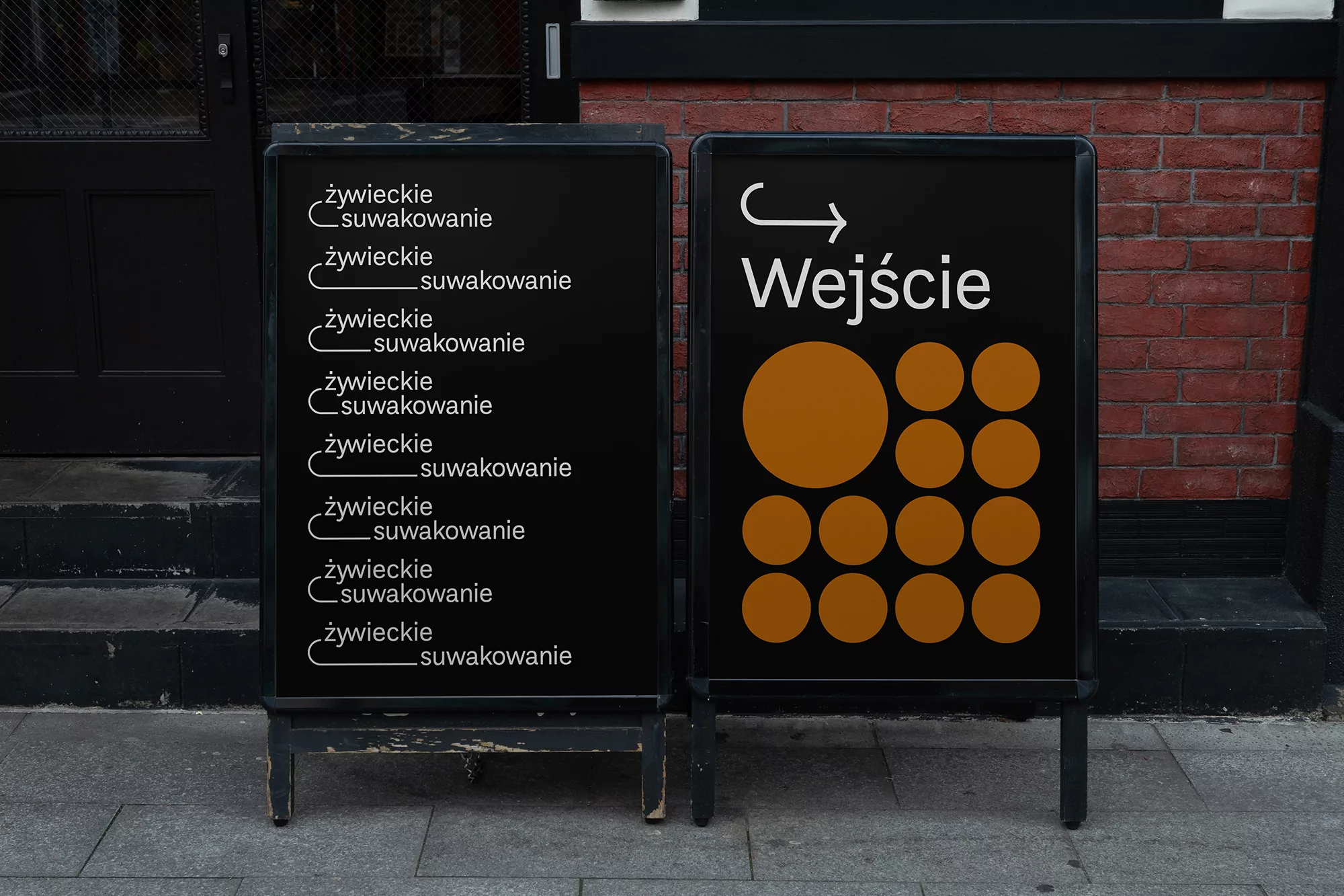 Two black sandwich boards with graphic designs and text in Polish on a city sidewalk with a brick building facade in the background.