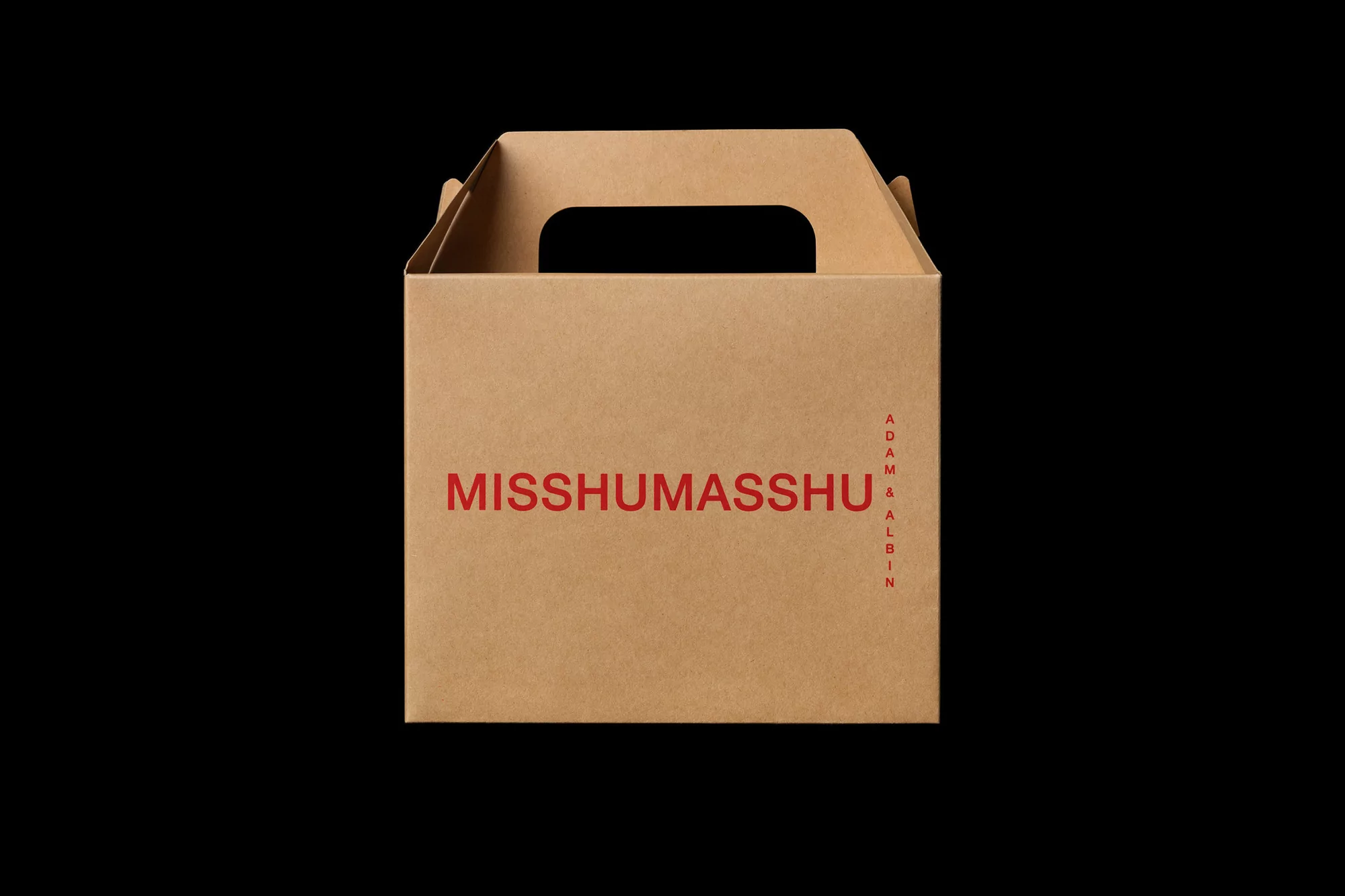 Brown cardboard box with the text 'MISSHUMASSHU' in red font and 'ADAM & ALBIN' vertically aligned. Isolated on black background.