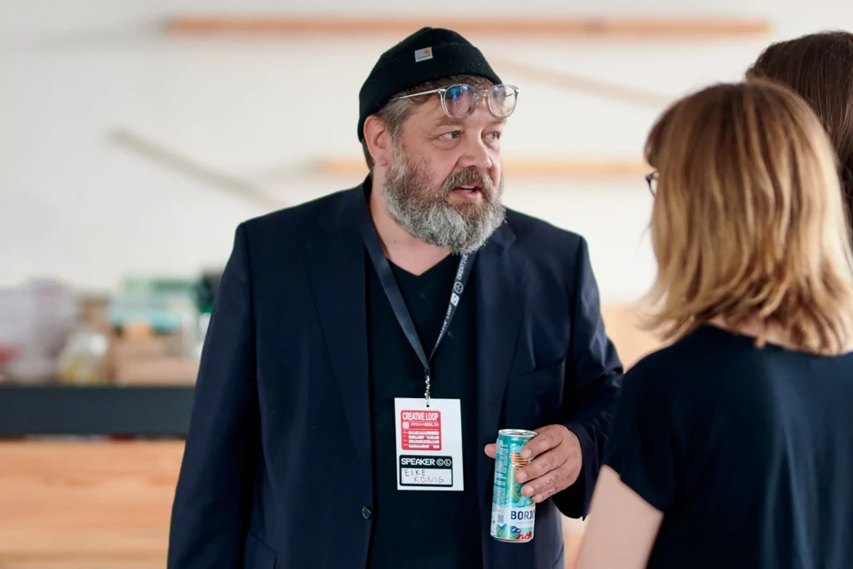 A man with a beard and glasses, wearing a black beanie and a blazer, stands indoors holding a drink while talking to someone.