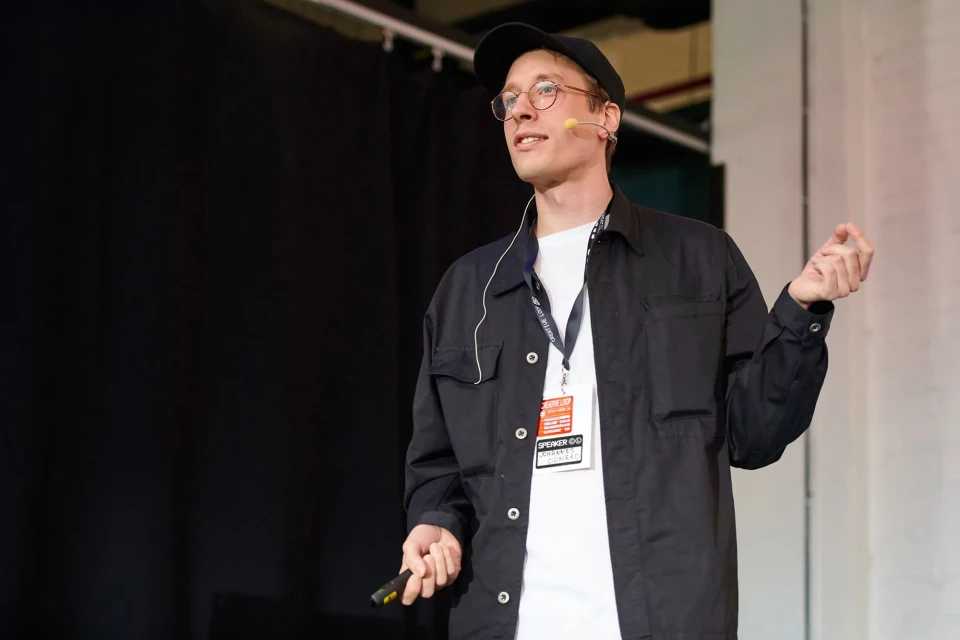 Adult male in a black jacket, white t-shirt, and cap speaks on stage with a lanyard and microphone against a dark backdrop.