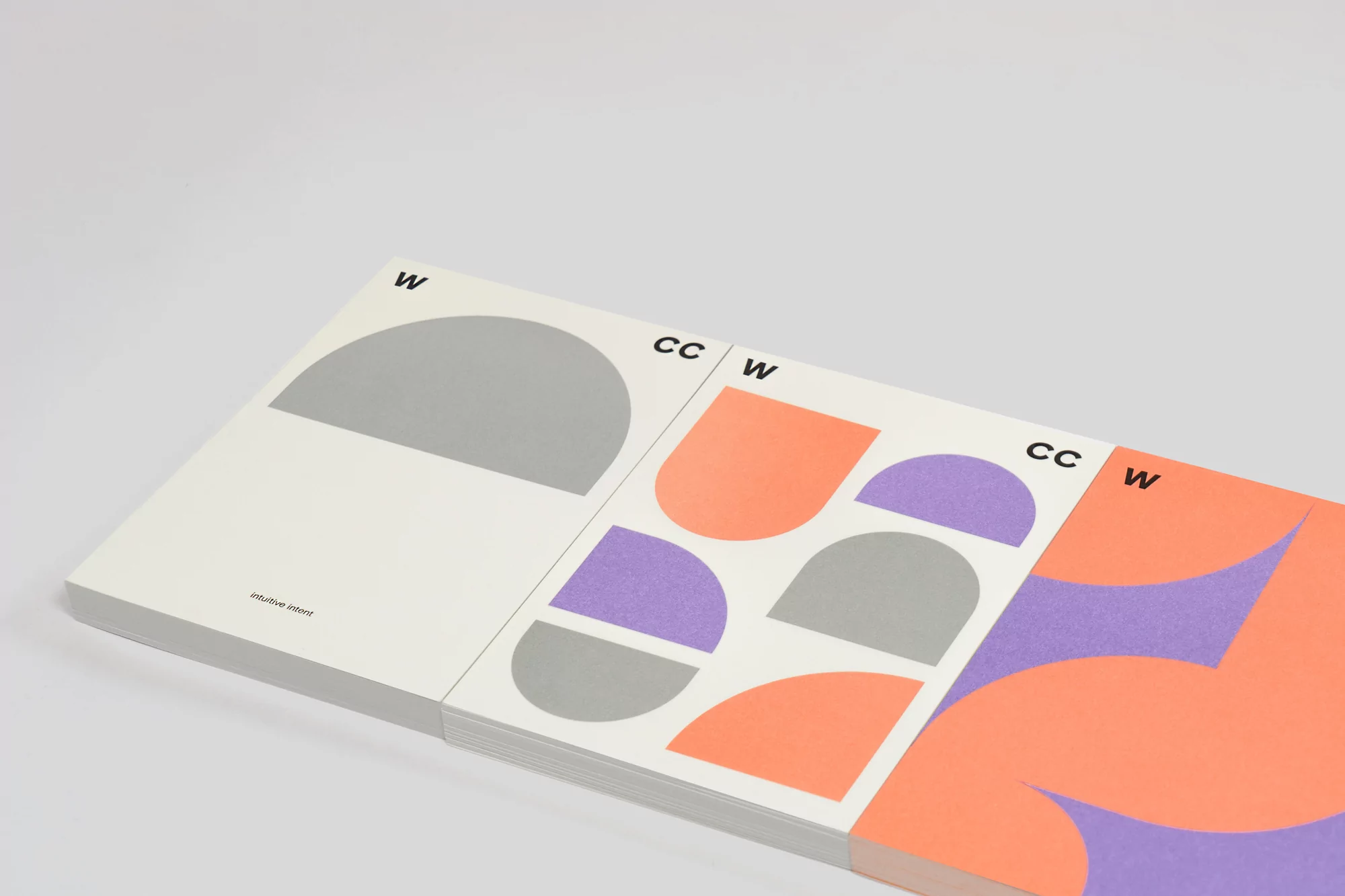 Stack of graphic design art books with geometric shapes in shades of gray, purple, and coral against a white background.
