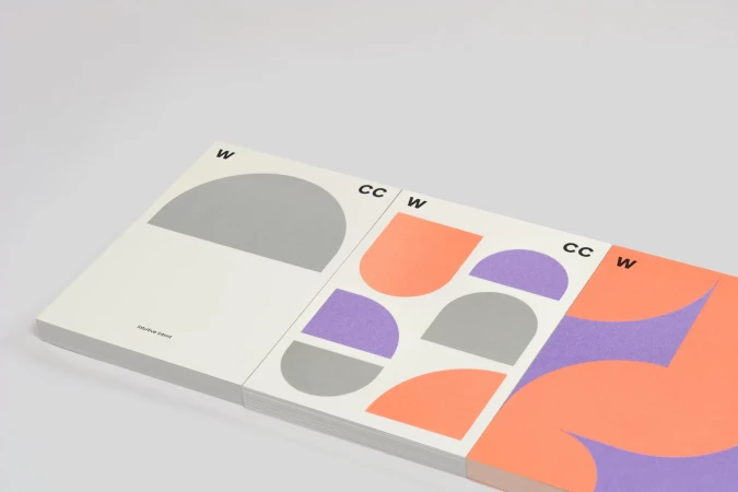 Stack of graphic design art books with geometric shapes in shades of gray, purple, and coral against a white background. 