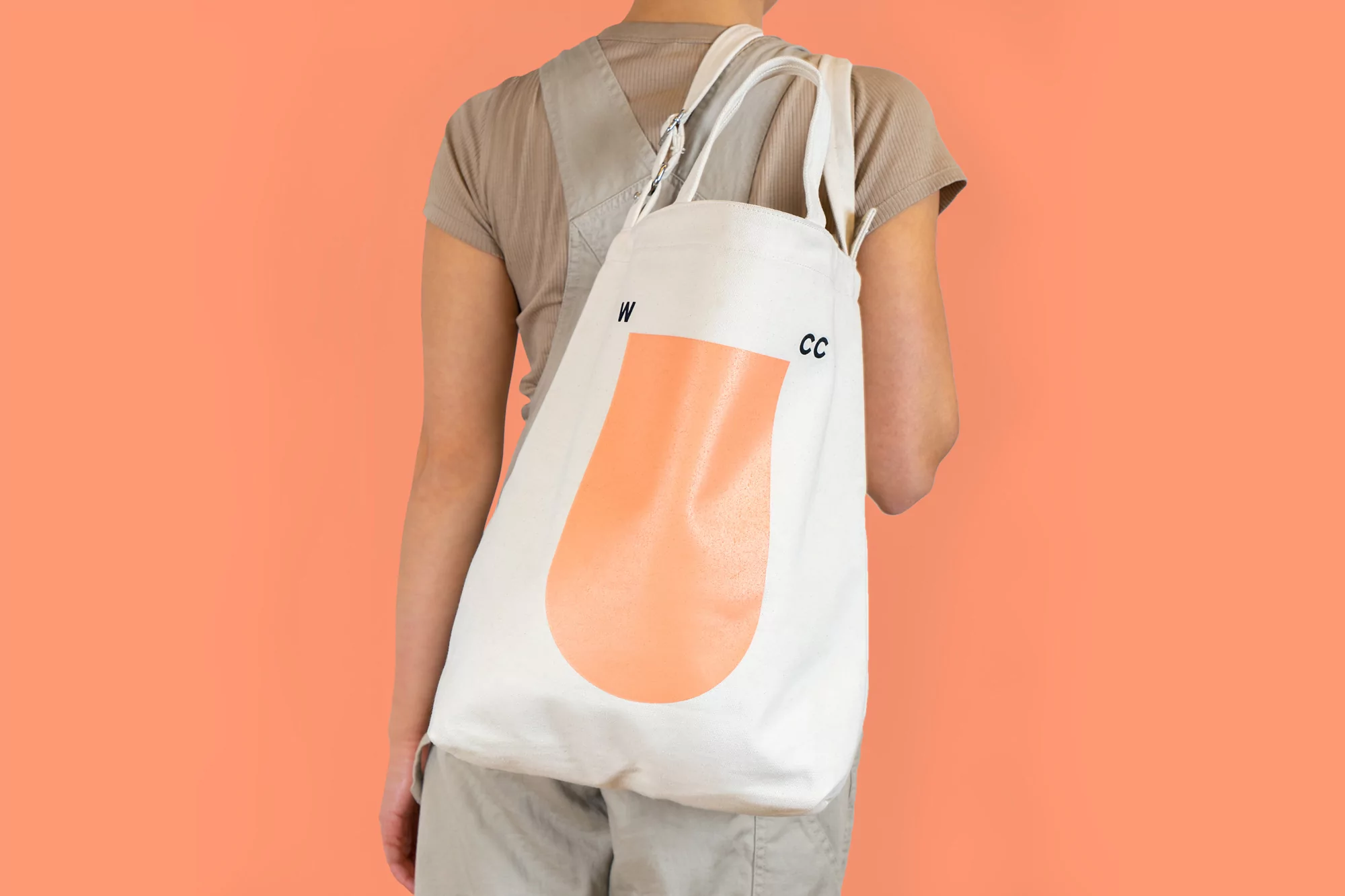 A person carries a white canvas tote bag with a peach-colored design against a peach background. Minimalist fashion and neutral colors.