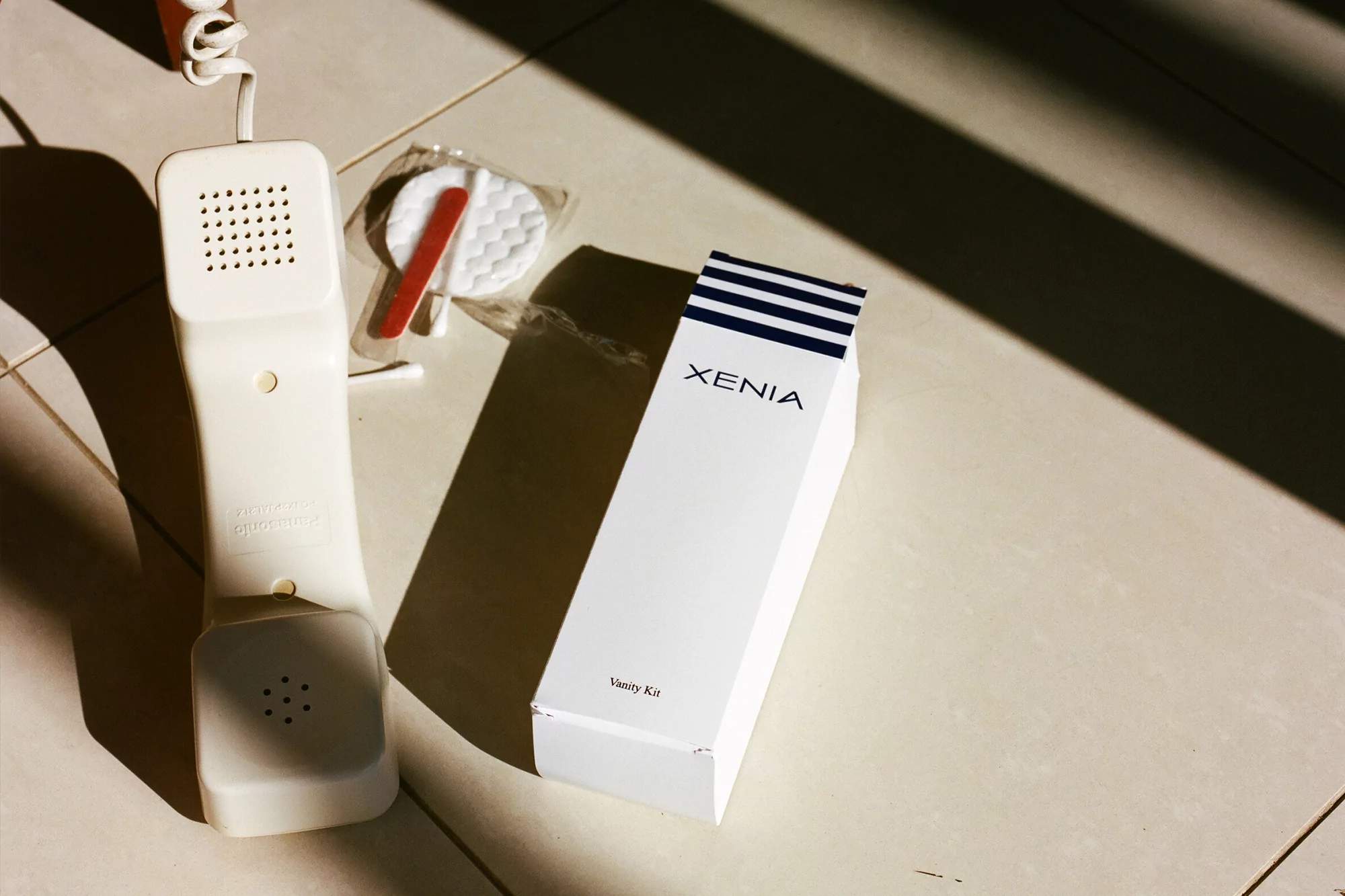 Still life of a telephone receiver, vanity kit, and cotton rounds on a tiled floor with shadows, set in a clean, minimal style.