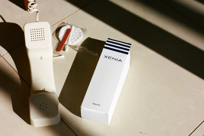 Still life of a telephone receiver, vanity kit, and cotton rounds on a tiled floor with shadows, set in a clean, minimal style.