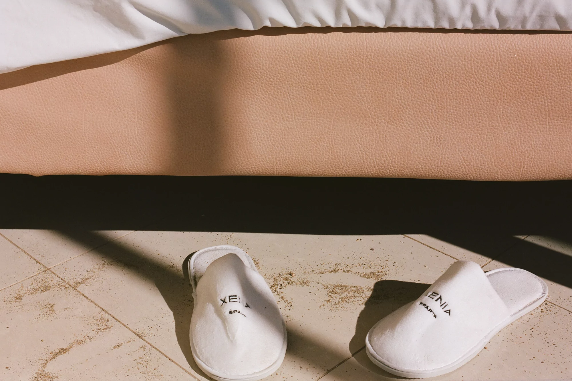 Close-up of a hotel room with two white spa slippers on a tiled floor, shadows, a bed base, and the corner of a white bed.