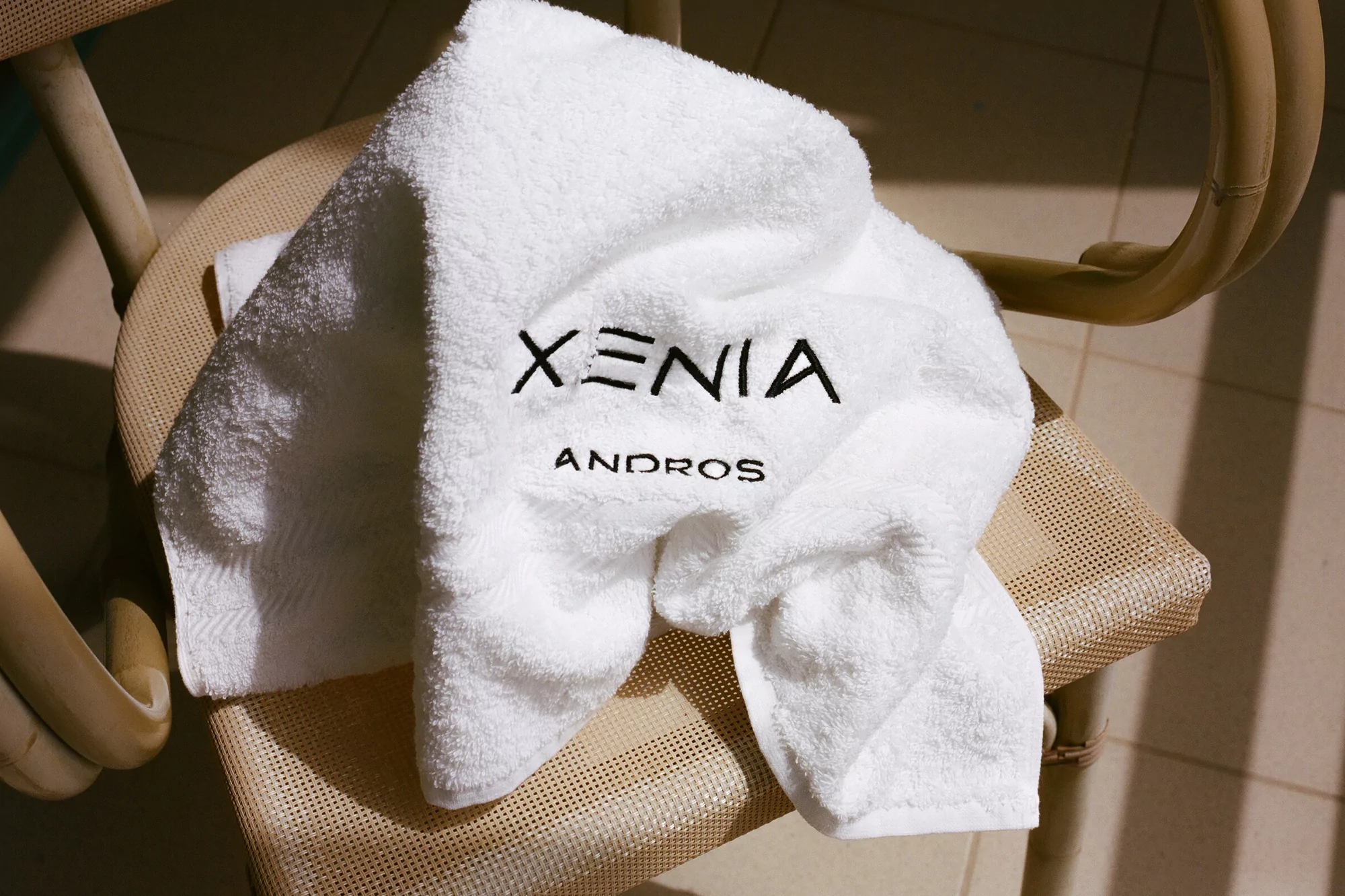 White towel with black embroidered text 'Xenia Andros' draped on a wicker chair in soft, neutral lighting.