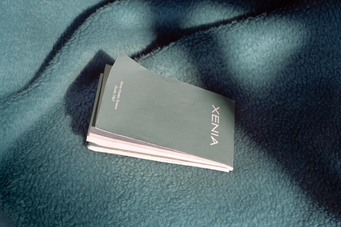 Stack of Xenia Hotels Greece Guide 1967 brochures, teal in color, are displayed on a teal blanket in a brightly lit environment.