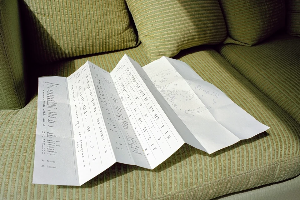 Folded white map with a text list sits on a green ridged couch with throw pillows, suggesting travel or trip planning.