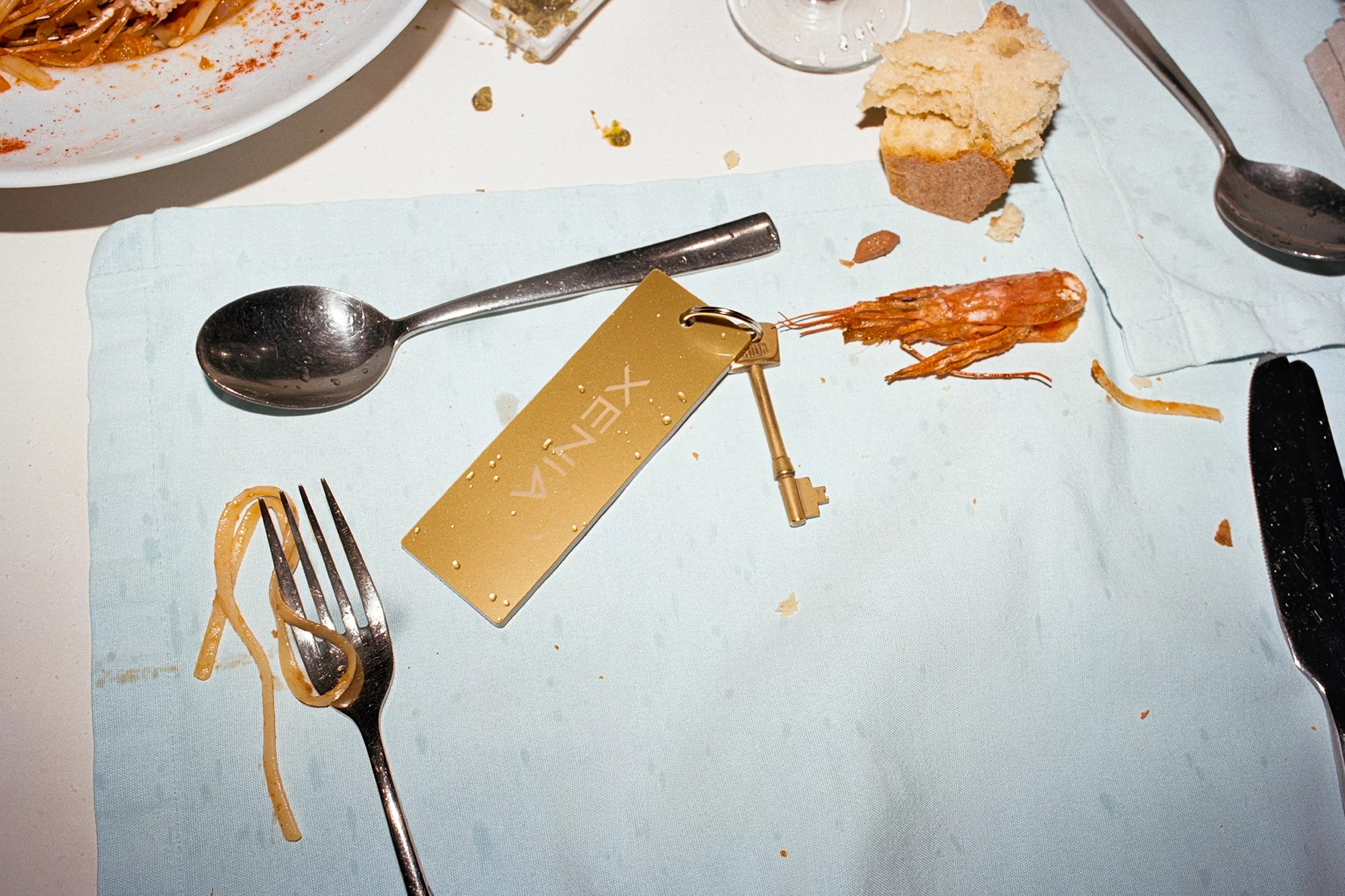 Overhead shot of place setting with fork, spoons, knife, plate, bread, key tag, and cooked shrimp on a light-blue tablecloth.