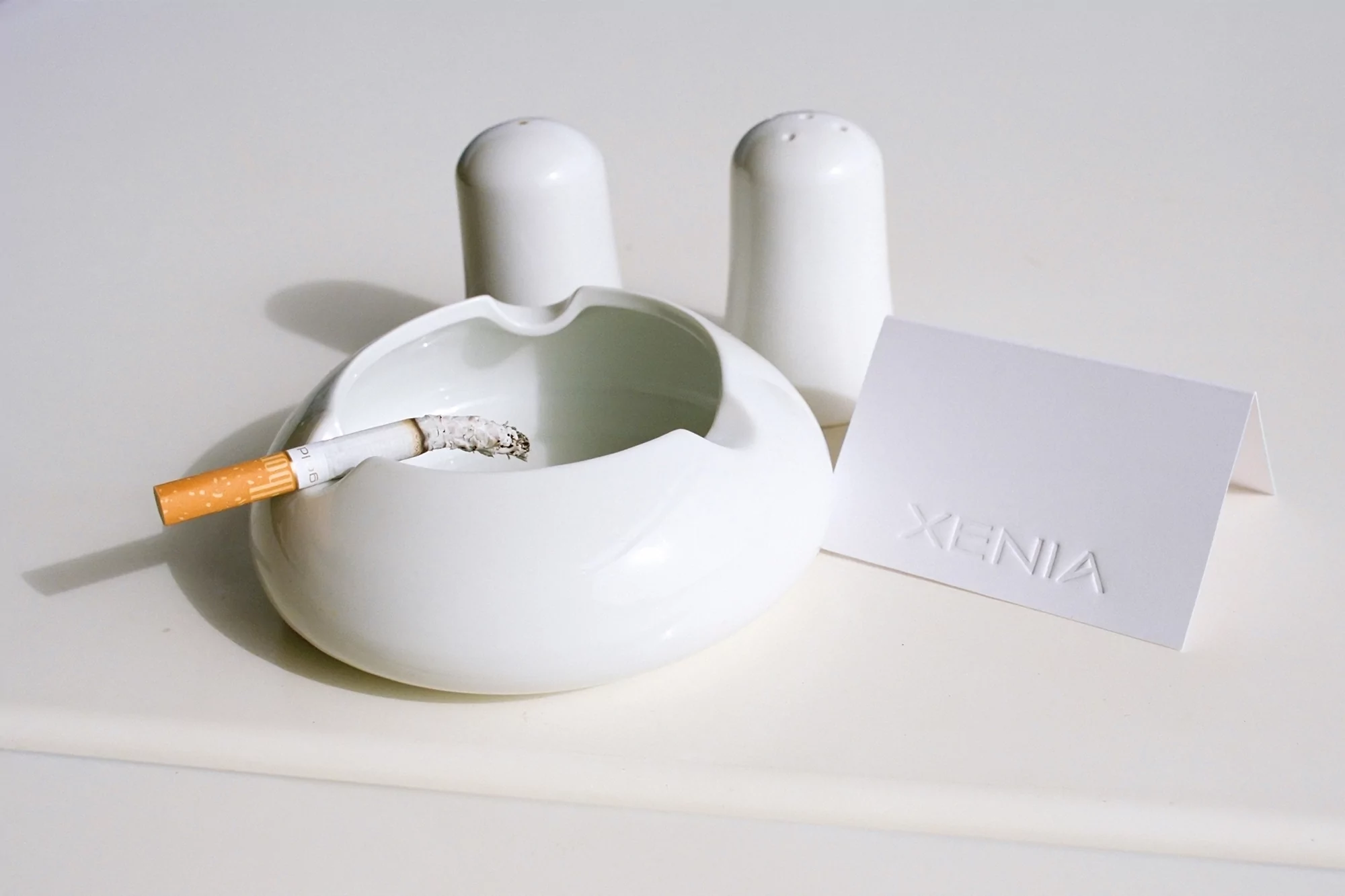 Still life of a lit cigarette in a white ashtray with salt and pepper shakers and a name card on a white surface.